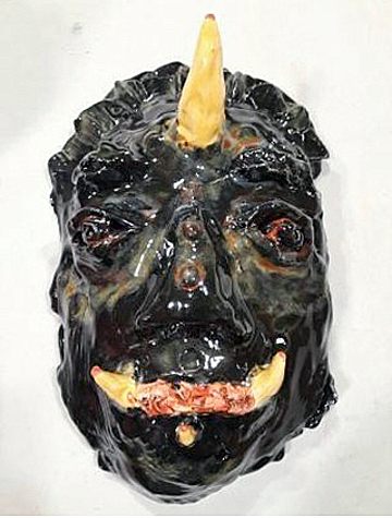 A glossy black ceramic mask depicting a mythical creature half human half dinosaur with orange horns