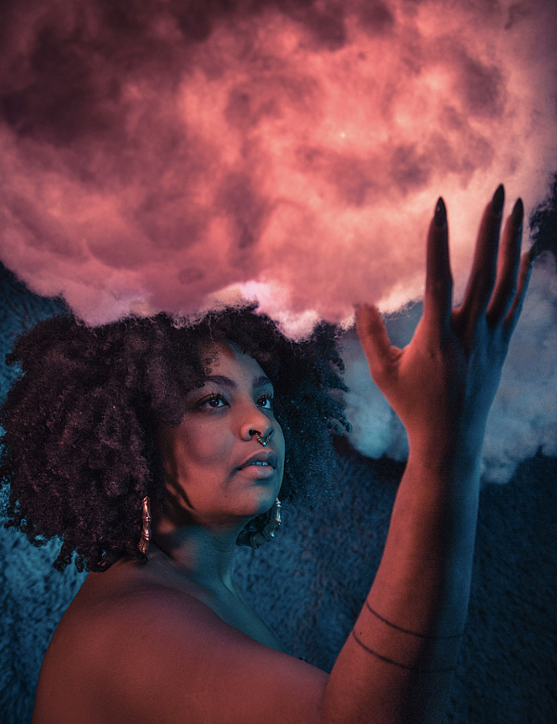 Color photo of Arielle gray, with her hand reaching towards a cloud above her head, which is backlit in shades of pink.