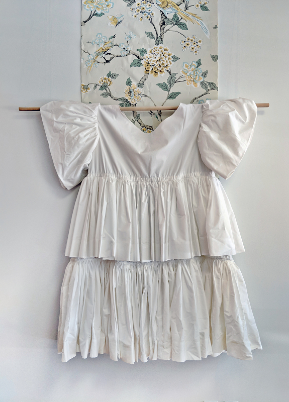 A white cotton dress with a wide boat neck, puffy sleeves and layered double skirt hangs from a pole, with a strip of floral wallpaper behind.