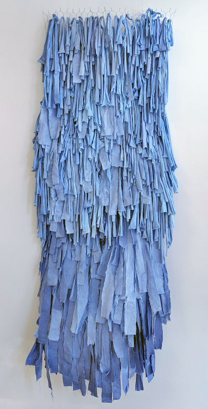 A sculptural piece made of strips of fabric died in different shades of blue, tied to a background made of chicken wire. The piece is titled \