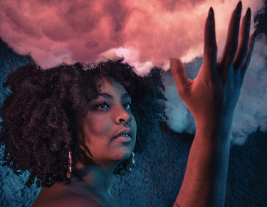 Color photo of Arielle gray, with her hand reaching towards a cloud above her head, which is backlit in shades of pink.