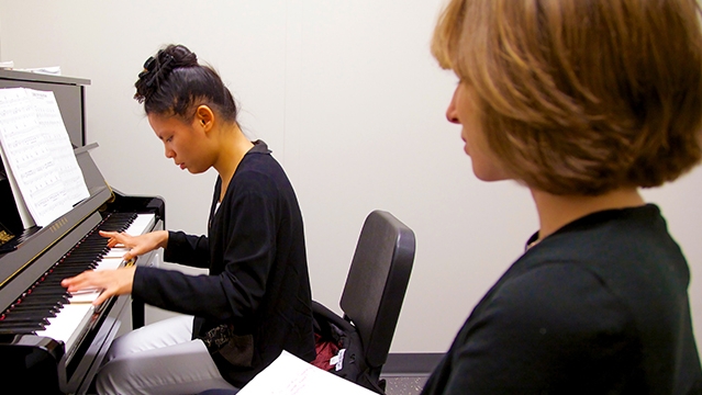 Woman playing piano with teacher watching class=